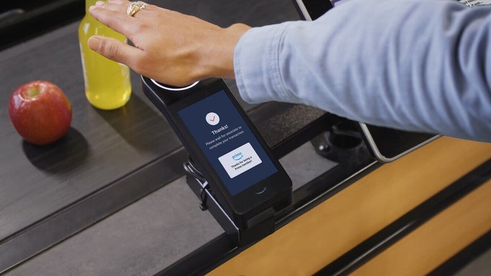 Human arm wearing a blue shirt holding the palm over an electric scanning device at a grocery store check-out lane.