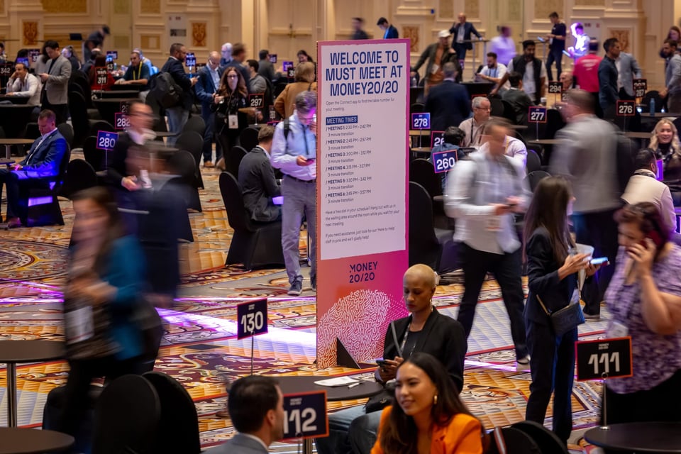 Image of a showroom floor, with people walking around and sitting at tables. A sign in the center reads "Welcome to must meet at Money20/20"