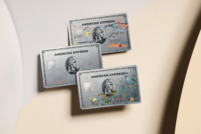 Three Platinum American Express credit cards, stacked above each other. The top card has multi-colored lines and stripes across it, the middle card is just platinum with no pattern, and the bottom has plants and flowers in the bottom right corner that spans across half of the card.