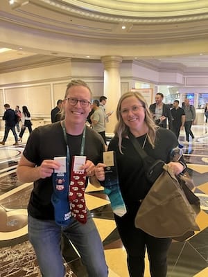 An image of a man and a woman holding three pairs of socks. The man is holding two pairs, one blue pair that says "buy low sell high", one that says "BaaS" with sheep. The woman is holding a pair of blue gradient socks, with the "Astra" logo.