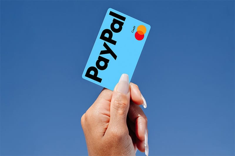 Woman with long white nails holding a light blue credit cad with the PayPal logo horizontally over the top of the card.