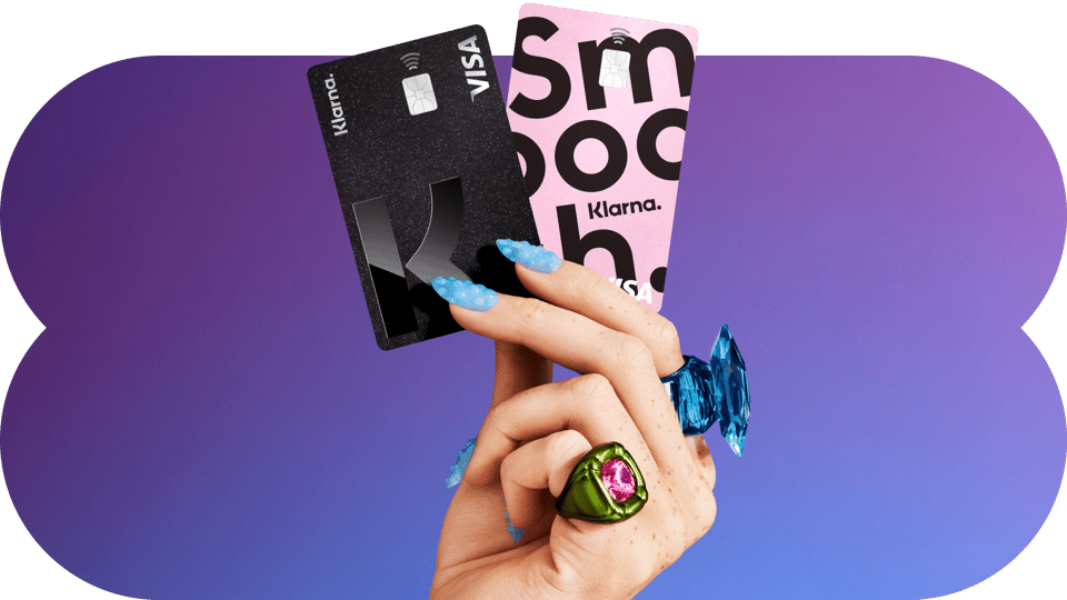 A woman's hand with bright blue finger nails and very large rings holds two credit cards, one black and one pick with large black lettering, for the company Klarna.