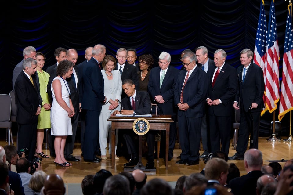 A group of men in dark suits and a few women in light colors watch President Barack Obama sign a document at a small table.