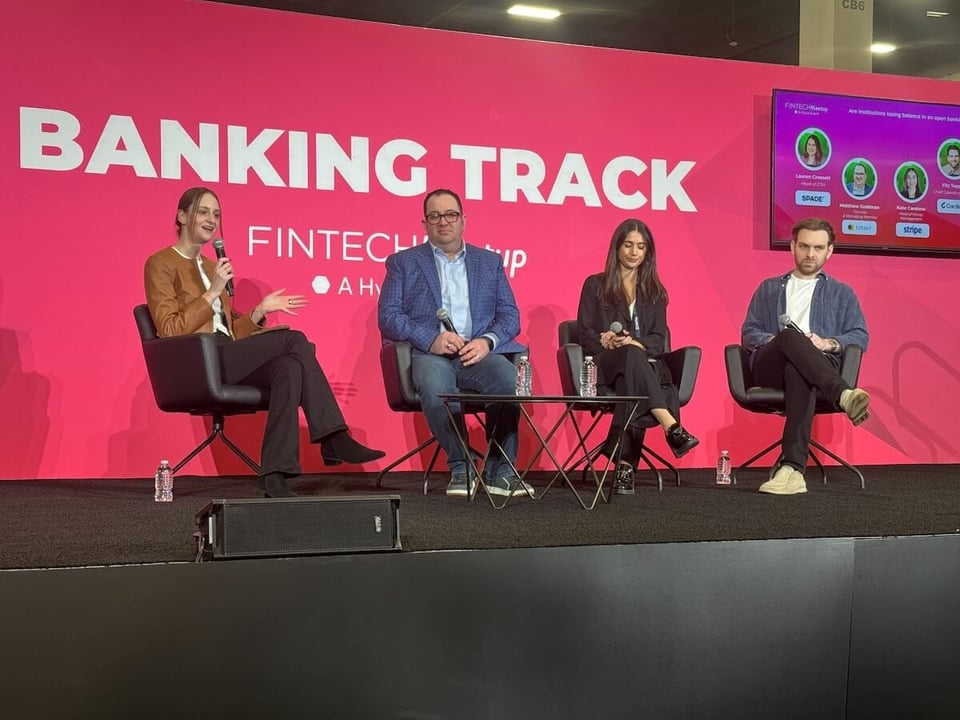 Photo of four people in business casual sitting on a black stage with a bright pink background, featuring the words "Banking Track" in white. Two of the people are men, two are women. No one looks particularly excited to be there.